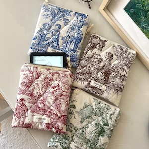 May include: Four quilted fabric pouches with zipper closures, each featuring a distinct toile de jouy print in blue, brown, red, or green. Each pouch has a small white label reading cutie pie. One red pouch contains a small electronic device.