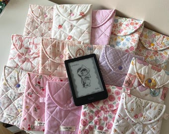 Puede incluir: Una colección de fundas de tela acolchada para lectores electrónicos, con varios estampados florales, de cuadros y rayas en tonos pastel con cierres a presión. Cada funda tiene una etiqueta que dice cutie pie. Un lector Kobo negro descansa encima.