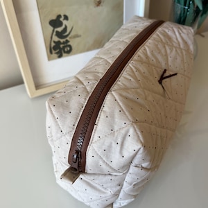 Pu&ograve; includere: Un beauty case trapuntato beige con cerniera marrone e piccoli pois neri. La borsa ha una forma rettangolare e un piccolo passante in tessuto marrone attaccato al lato. La borsa &egrave; chiusa.