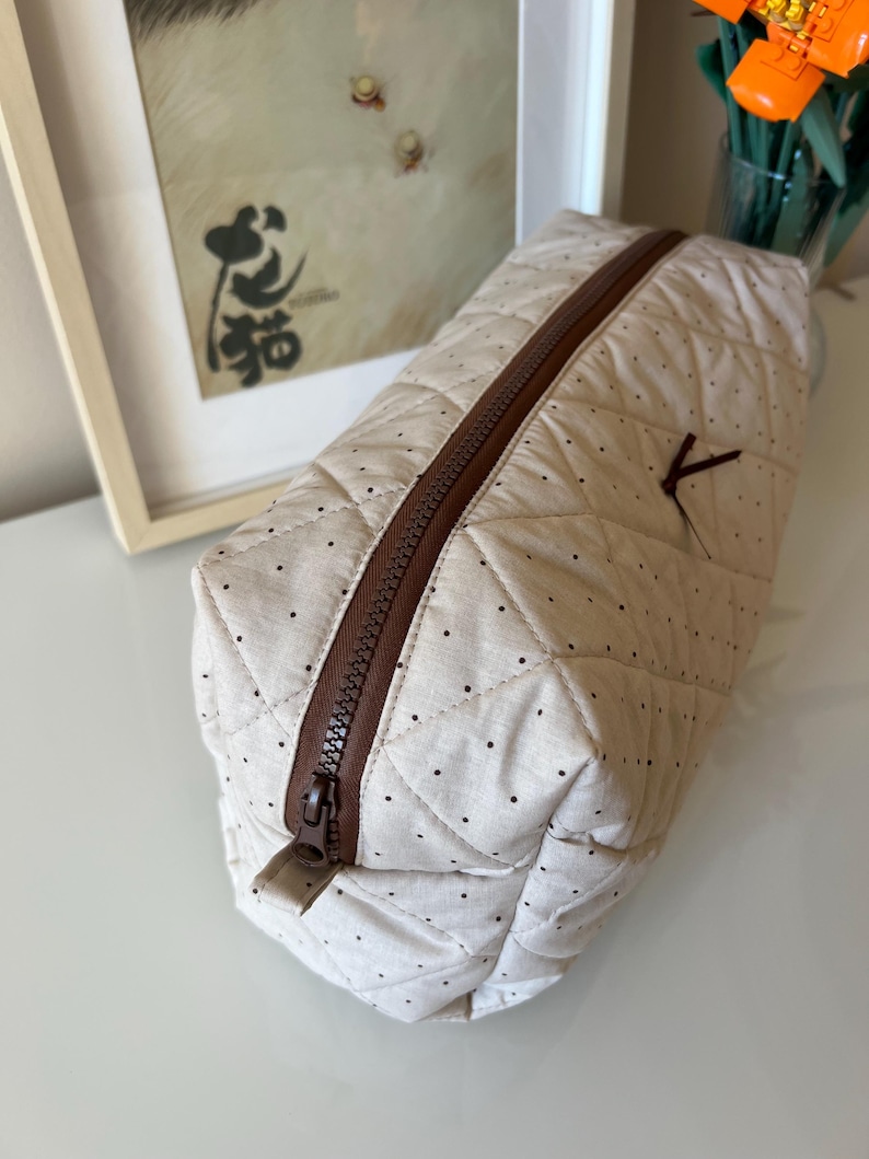 Pu&ograve; includere: Un beauty case trapuntato beige con cerniera marrone e piccoli pois neri. La borsa ha una forma rettangolare e un piccolo passante in tessuto marrone attaccato al lato. La borsa &egrave; chiusa.