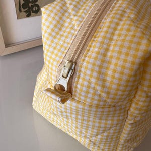 May include: A yellow and white gingham patterned cosmetic bag with a beige zipper. The bag has a quilted design and a rectangular shape. The zipper is open, revealing the interior. The bag is sitting on a white surface.