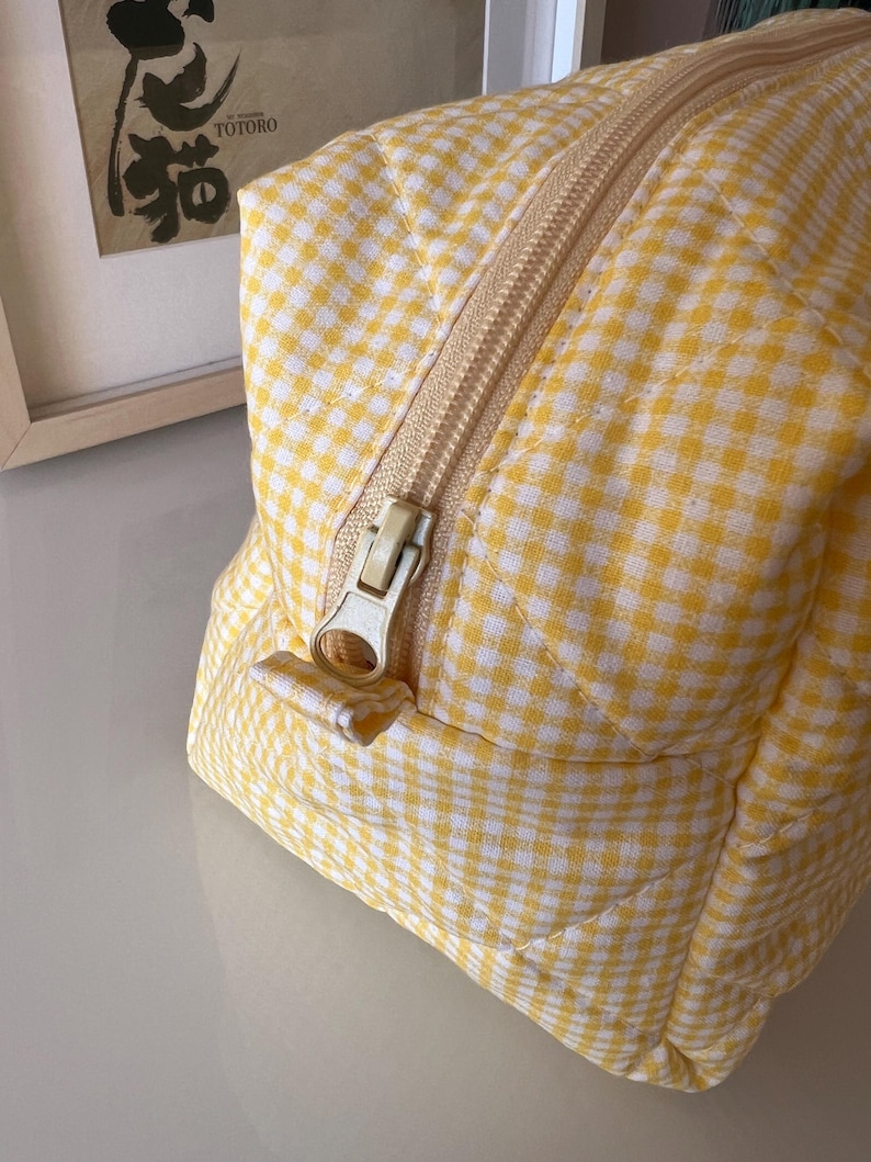 May include: A yellow and white gingham patterned cosmetic bag with a beige zipper. The bag has a quilted design and a rectangular shape. The zipper is open, revealing the interior. The bag is sitting on a white surface.