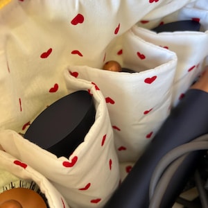 May include: A white cosmetic bag with red heart patterns, featuring multiple pockets. The bag contains a hair dryer, a hairbrush, and other beauty accessories. The interior is lined with a white fabric with red heart shapes.