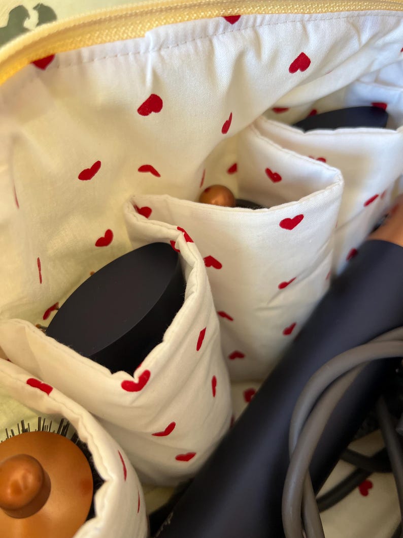 May include: A white cosmetic bag with red heart patterns, featuring multiple pockets. The bag contains a hair dryer, a hairbrush, and other beauty accessories. The interior is lined with a white fabric with red heart shapes.
