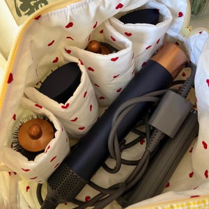 May include: Open yellow cosmetic bag with a gingham exterior and a white interior patterned with red hearts. The bag contains a dark blue hair dryer, brushes, and other beauty tools. The bag is open, revealing the contents.