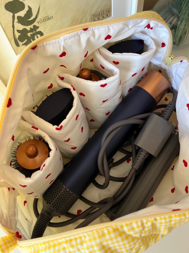 May include: Open yellow cosmetic bag with a gingham exterior and a white interior patterned with red hearts. The bag contains a dark blue hair dryer, brushes, and other beauty tools. The bag is open, revealing the contents.