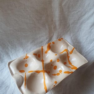 Könnte beinhalten: Ein rechteckiges Eierablage-Tablett aus Keramik mit einer glänzenden cremefarbenen Glasur und einem handgemalten orangefarbenen Spritzer- und Tropfenmuster. Das Tablett verfügt über sechs abgerundete Vertiefungen in einer zwei-mal-drei-Anordnung.