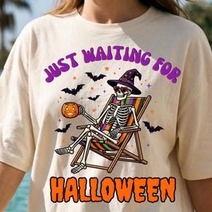 May include: Beige t-shirt with a Halloween-themed graphic. The design features a skeleton wearing sunglasses and a witch hat, relaxing in a beach chair, holding a jack-o'-lantern, and surrounded by bats. The text reads "JUST WAITING FOR HALLOWEEN".