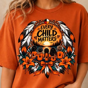 May include: An orange t-shirt featuring a graphic design with the text "EVERY CHILD MATTERS" above a sunset landscape. The illustration includes five children with their arms around each other, framed by feathers, orange flowers, and a bow.
