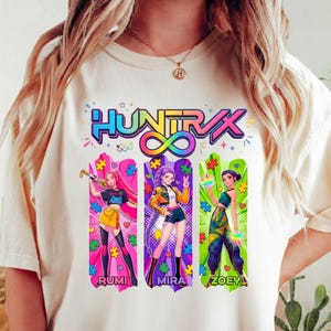 May include: A cream-coloured t-shirt with a vibrant graphic of three anime-style figures. The design includes the text "HUNTRVX" above the figures and the names "RUMI", "MIRA", and "ZOEY" below. The graphic uses bright colours and puzzle piece details.