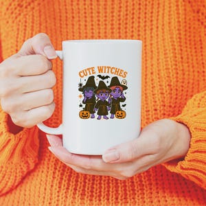 May include: White ceramic mug with the words "Cute Witches" and a graphic of three cartoon witches in black hats and robes, with pumpkins. The mug is held by a person wearing an orange jumper.