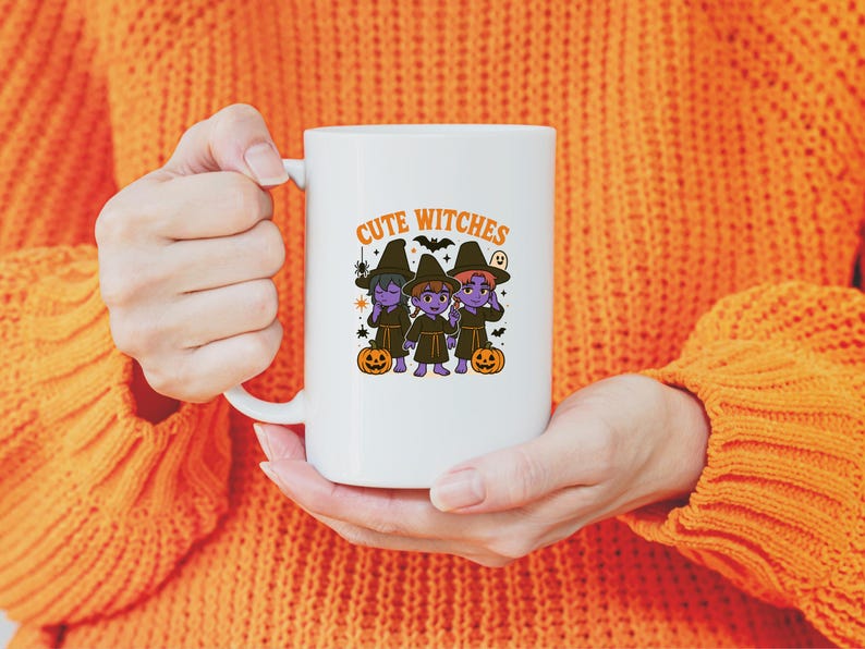 May include: White ceramic mug with the words "Cute Witches" and a graphic of three cartoon witches in black hats and robes, with pumpkins. The mug is held by a person wearing an orange jumper.