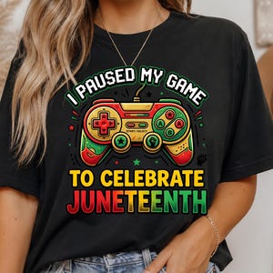 Op de afbeelding: Zwart T-shirt met een kleurrijk ontwerp van een videogamecontroller in rood, groen en geel. De tekst luidt: "IK PAUZERDE MIJN SPEL OM JUNETEENTH TE VIEREN."