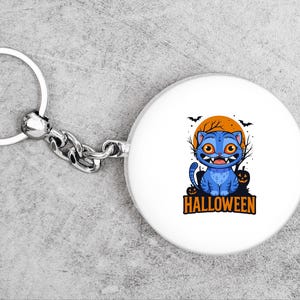 May include: A white keychain with a round charm featuring a blue cartoon cat with orange eyes and sharp teeth. The cat is set against an orange moon with bats and bare trees. The word "HALLOWEEN" is below the cat.