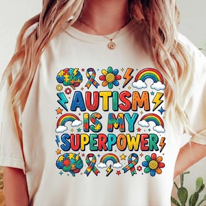 May include: Cream-colored t-shirt with the text "AUTISM IS MY SUPERPOWER" in colorful, bold letters. The design includes rainbows, puzzle pieces, lightning bolts, and flowers, all in vibrant colors. The shirt is a casual, everyday wear item.