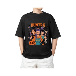 May include: Black t-shirt with a Halloween graphic, featuring the word "HUNTRX" in orange. The design includes three cartoon figures, pumpkins, a black cat, and other spooky elements. The shirt is worn by a person.