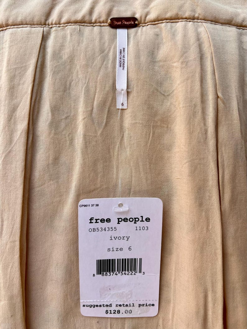 May include: Close-up of a cream-colored garment with a tag from Free People. The tag indicates the size is 6 and the color is ivory. The suggested retail price is $128.00.