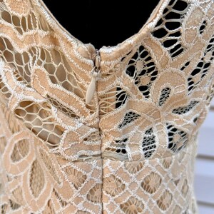 May include: Close-up of a beige lace dress with a V-neckline and a zipper closure. The intricate lace pattern features floral and geometric designs. The dress is made of a delicate, open-weave fabric, perfect for special occasions.
