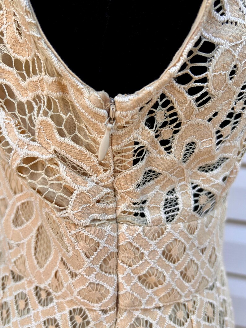 May include: Close-up of a beige lace dress with a V-neckline and a zipper closure. The intricate lace pattern features floral and geometric designs. The dress is made of a delicate, open-weave fabric, perfect for special occasions.