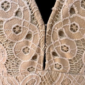 May include: Close-up of a beige lace garment with a plunging neckline. The intricate lace design features floral patterns and openwork details. The garment is fastened with a small clasp at the neckline.