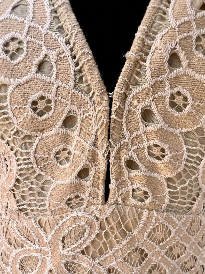 May include: Close-up of a beige lace garment with a plunging neckline. The intricate lace design features floral patterns and openwork details. The garment is fastened with a small clasp at the neckline.