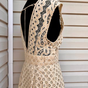 May include: A beige lace dress displayed on a black mannequin. The dress features a low back and a fitted waist. The lace has a detailed floral and geometric pattern. The dress is sleeveless and has a flowing skirt.