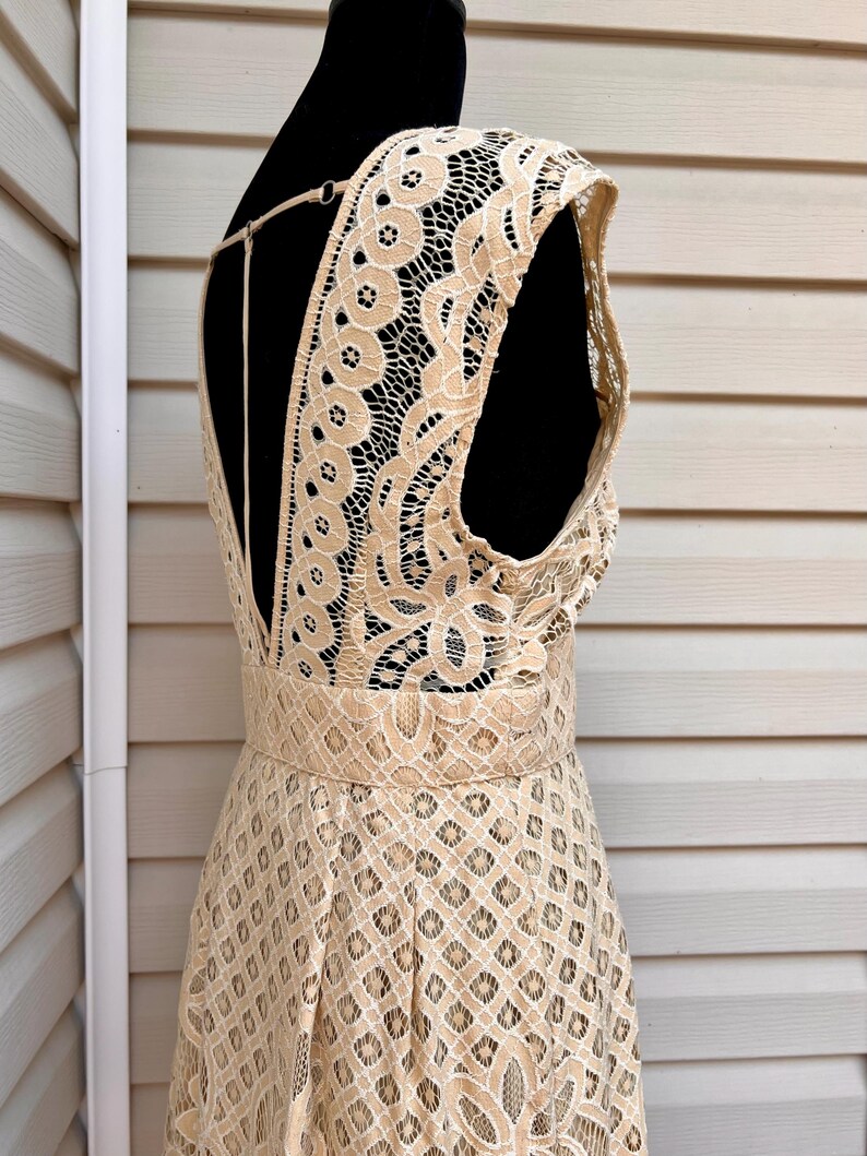 May include: A beige lace dress displayed on a black mannequin. The dress features a low back and a fitted waist. The lace has a detailed floral and geometric pattern. The dress is sleeveless and has a flowing skirt.