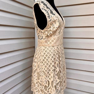 May include: A beige lace dress with a V-neckline and sleeveless design. The dress features a fitted waist and a flared skirt, showcasing intricate floral and geometric patterns. The dress is displayed on a black mannequin.