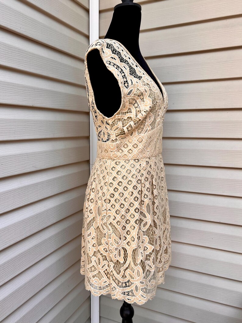 May include: A beige lace dress with a V-neckline and sleeveless design. The dress features a fitted waist and a flared skirt, showcasing intricate floral and geometric patterns. The dress is displayed on a black mannequin.