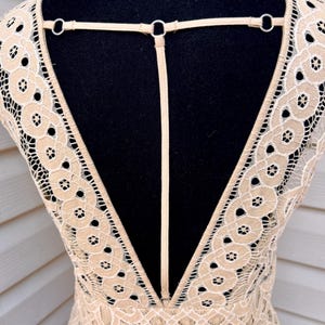 May include: A beige lace dress with a deep V-neck and open back design. The dress features intricate floral patterns and a delicate strap detail across the upper back, creating an elegant and airy look.
