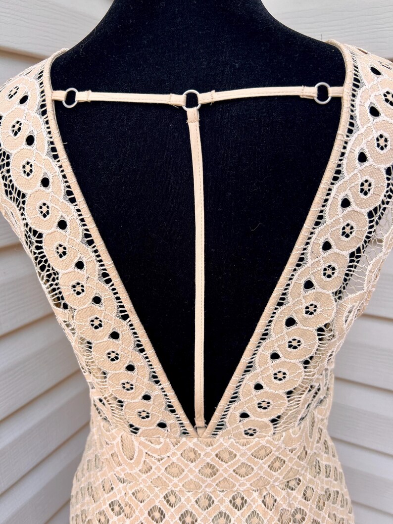 May include: A beige lace dress with a deep V-neck and open back design. The dress features intricate floral patterns and a delicate strap detail across the upper back, creating an elegant and airy look.