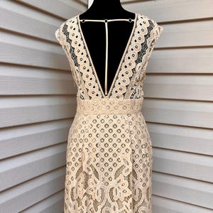 May include: A beige lace dress with a deep V-neck and open back detail. The dress features a fitted bodice and a flared skirt, with intricate floral and geometric patterns throughout the lace. The dress is displayed on a black mannequin.