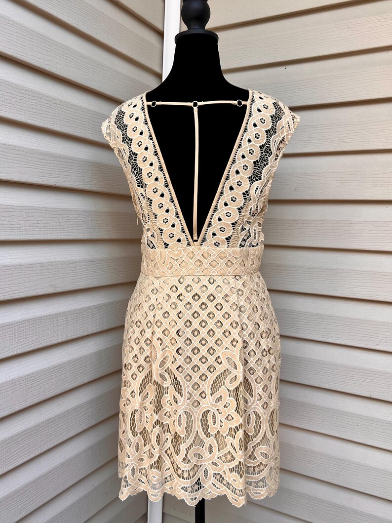 May include: A beige lace dress with a deep V-neck and open back detail. The dress features a fitted bodice and a flared skirt, with intricate floral and geometric patterns throughout the lace. The dress is displayed on a black mannequin.