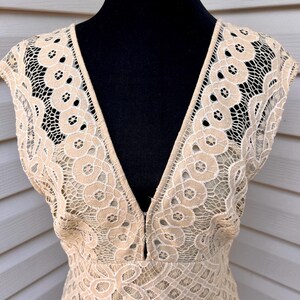 May include: A beige lace dress with a deep V-neckline. The dress features a delicate floral lace pattern with a scalloped edge on the sleeves. The dress is displayed on a black mannequin.
