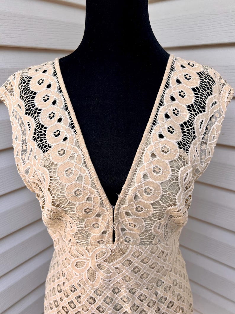 May include: A beige lace dress with a deep V-neckline. The dress features a delicate floral lace pattern with a scalloped edge on the sleeves. The dress is displayed on a black mannequin.