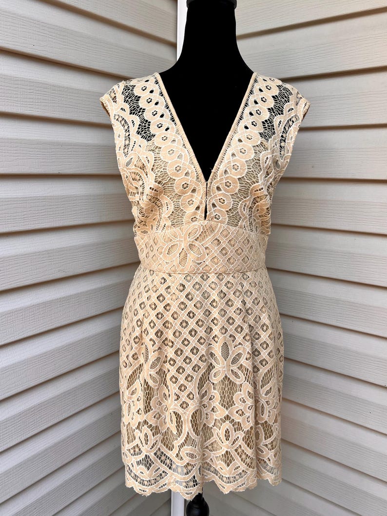 May include: A beige lace dress with a V-neck and cap sleeves. The dress features a fitted waist and a flared skirt. The lace has a floral and geometric pattern. The dress is displayed on a black mannequin.