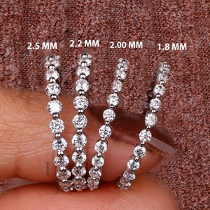 Könnte beinhalten: Vier silberne Ringe mit klaren Edelsteinen werden gezeigt. Die Ringe sind mit Größen beschriftet: 2,5 mm, 2,2 mm, 2,00 mm und 1,8 mm. Die Ringe werden vor einem braunen Hintergrund gehalten.