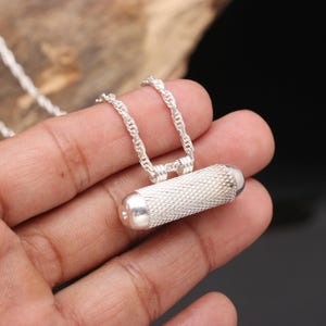 Puede incluir: Un collar con colgante de plata con un dije cilíndrico texturizado con extremos redondeados. El dije cuelga de una delicada cadena de plata. La superficie del colgante tiene un patrón detallado de trama cruzada.
