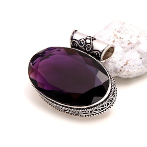May include: A large, oval-shaped pendant with a deep purple gemstone. The gemstone is set in a silver-colored metal frame with intricate detailing. The pendant has a loop at the top for a chain. The background is white.