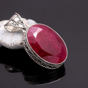 May include: An oval pendant featuring a large, faceted, deep red gemstone. The gemstone is set in a silver-coloured metal with an ornate, filigree bail. The pendant is displayed on a white, textured surface against a dark background.