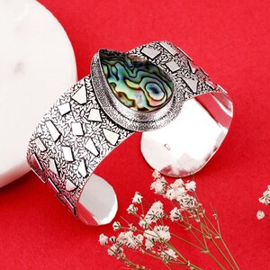 May include: Silver cuff bracelet with an oval abalone shell centrepiece displaying iridescent green, blue, and purple hues. The bracelet has a textured, geometric pattern and a wide, open design.