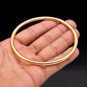 May include: A gold-colored bangle bracelet. The circular bracelet is smooth and shiny, with a simple, elegant design. The bangle is held in a person's hand against a black background.