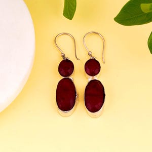 Pode incluir: Um par de brincos pendurados em prata com duas pedras preciosas vermelhas cada. A pedra superior é redonda e a inferior oval. Os brincos são exibidos em um fundo amarelo.