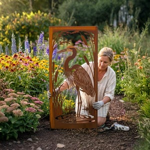 May include: A rectangular, rust-colored metal garden screen featuring a heron design. The heron is depicted amidst cattails and water ripples. The screen is set in a garden with colorful flowers.