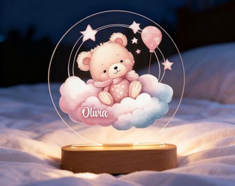 May include: A night light featuring a teddy bear design. The bear is pink and seated on a cloud, with a pink balloon and stars. The name "Olivia" is written on the cloud. The light sits on a wooden base.