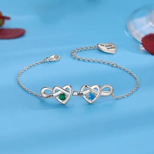 Op de afbeelding: Zilveren armband met twee oneindige hartbedels, elk met een edelsteen. Eén hart heeft een groene steen en de naam "Loana", de andere heeft een blauwe steen en de naam "Max". Een kleine hartbedel met de tekst "Diese Mutter" is ook inbegrepen.