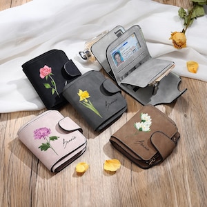 May include: A collection of small, floral-decorated wallets in various colors, including black, gray, and pink. Each wallet features a different flower design, such as a rose, daffodil, and chrysanthemum. Some wallets are open, revealing card slots and a clear ID window.