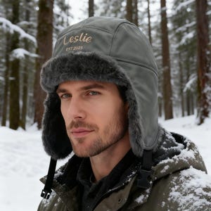 Può includere: Un cappello invernale grigio con paraorecchie e finiture in ecopelliccia. Il cappello ha il nome "Leslie" e l'anno "EST 2025" ricamati sul davanti. Il cappello è indossato in un ambiente di foresta innevata.
