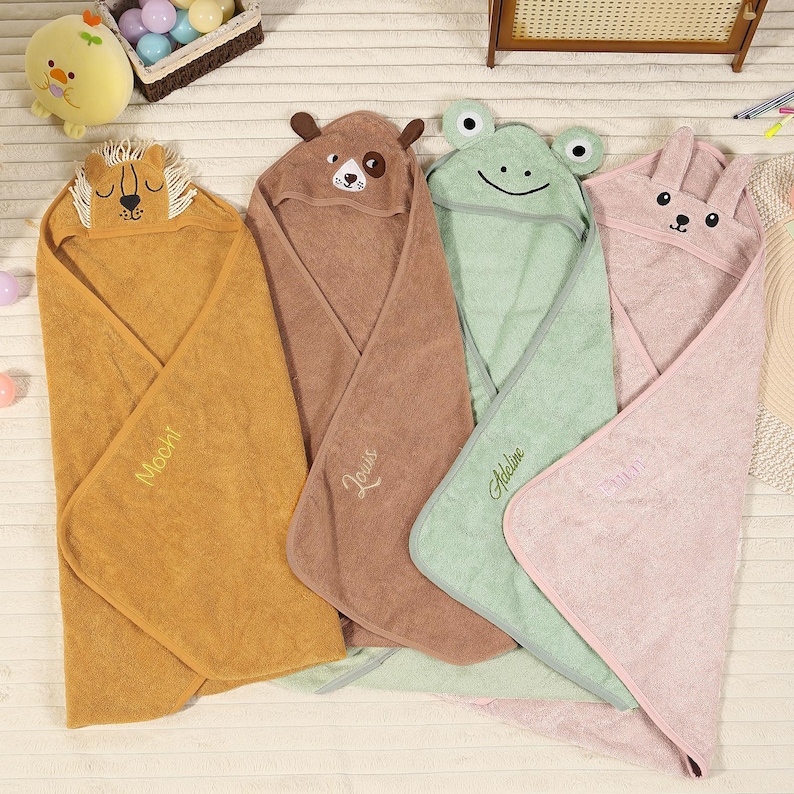 Pode incluir: Quatro toalhas com capuz para beb&ecirc;s em tons de dourado, marrom, verde e rosa. Cada toalha apresenta um rosto de animal no capuz: um le&atilde;o, um cachorro, um sapo e um coelho. Os nomes "Mochi", "Louis" e "Adeline" s&atilde;o bordados nas toalhas.
