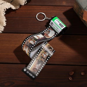 May include: A personalised film roll keyring featuring a green and black film canister and a strip of printed photos. The keyring includes a silver ring and a black and white film strip with multiple photos.